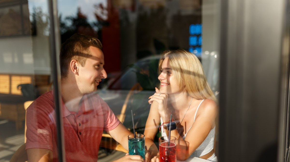 Mann und Frau auf einem Date mit zwei Drinks, hinter einer Glasscheibe, die sich gegenseitig anlächeln