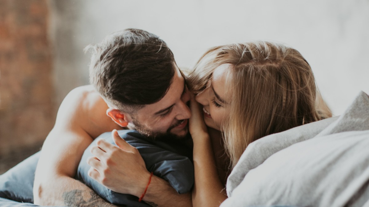 Frau und Mann im Bett zwischen Kissen, die sich anlächeln und umarmen