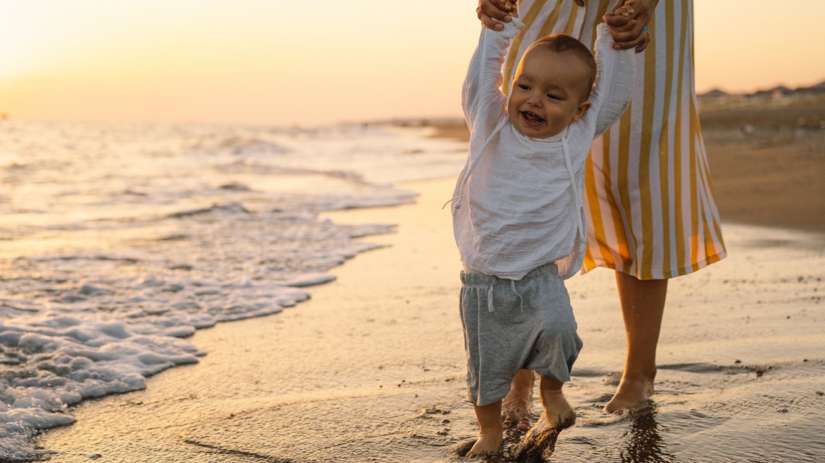 Frau mit Baby am Strand, das durch den Sand am Wasser geht und sich mit den Armen an der Mutter festhält im Sonnenuntergang