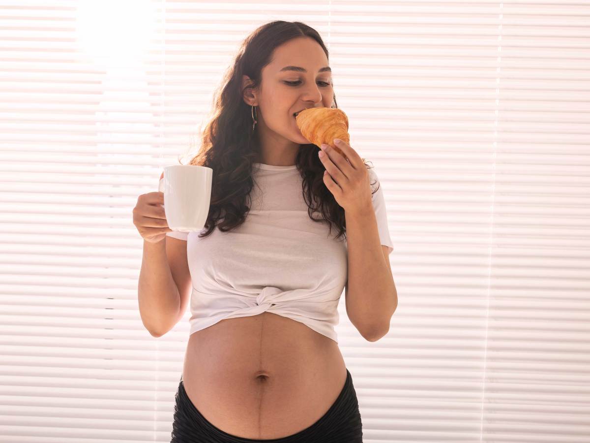 Schwangere Frau hält eine Tasse in der Hand und beißt genüsslich in ein Croissant.