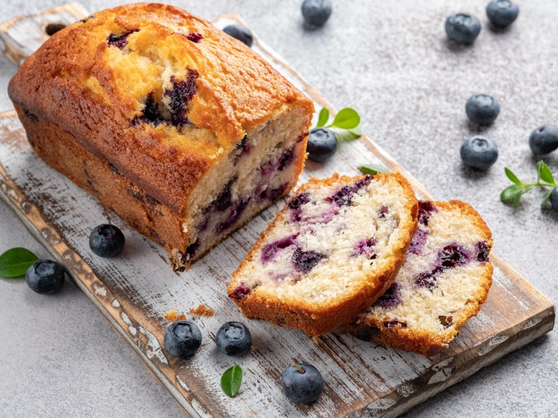 Ein angeschnittener Blaubeer-Zitronenkuchen auf einem Brett, zwei Scheiben abgeschnitten, drumherum frische Blaubeeren.