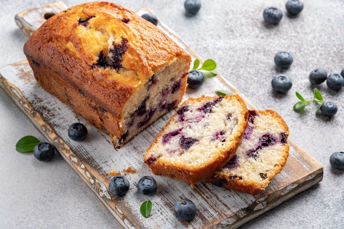 Ein angeschnittener Blaubeer-Zitronenkuchen auf einem Brett, zwei Scheiben abgeschnitten, drumherum frische Blaubeeren.