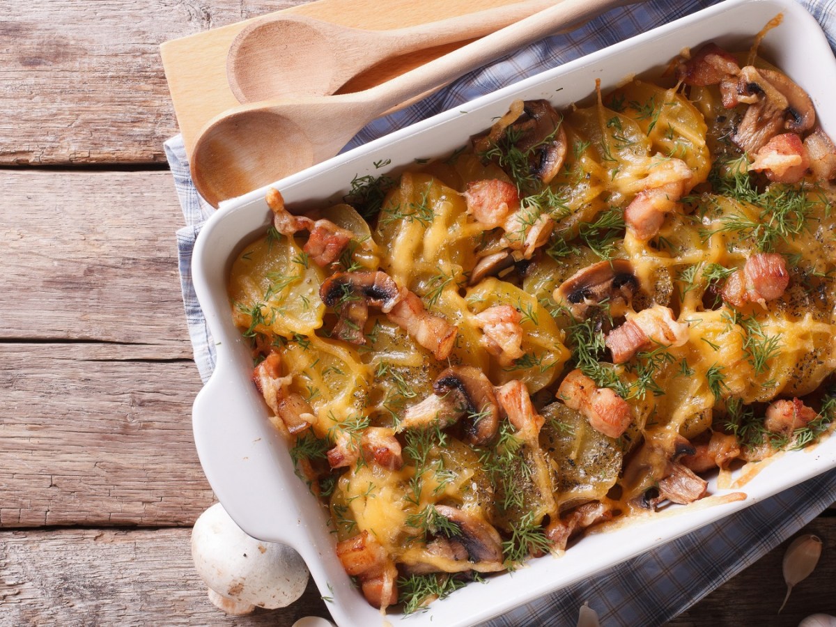 Eine weiße Auflaufform auf einem hellen Holztisch mit dem Gratin und einem Bund Kräuter sowie Pilzen und Knoblauchknollen.