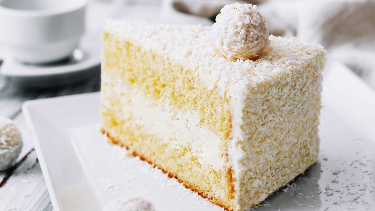 Ein Stück vom weißen Raffaello-Kuchen auf einem weißen Teller und hellem Holztisch.