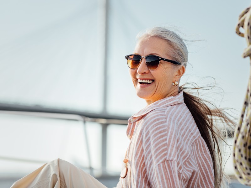 Frau über 50 sitzt draußen und trägt einen Pferdeschwanz und eine Sonnenbrille.