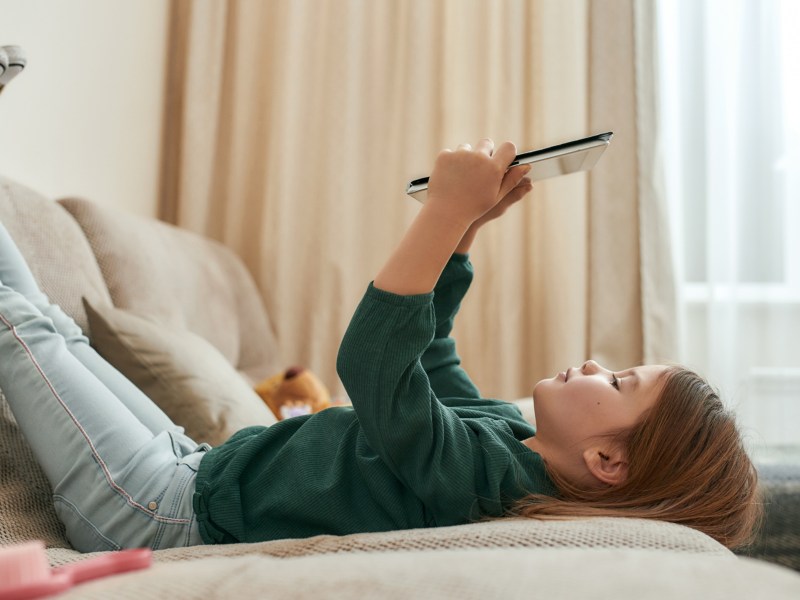 Kind, dass auf der Couch liegt und ein Tablet in der Hand hält und draufschaut