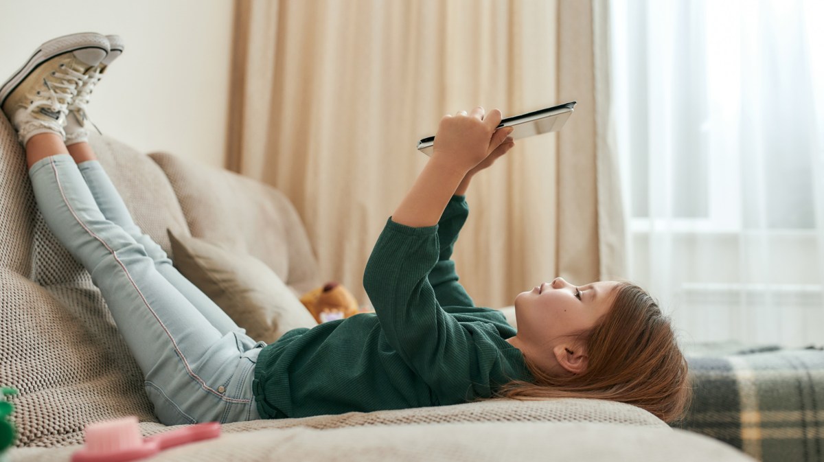 Kind, dass auf der Couch liegt und ein Tablet in der Hand hält und draufschaut