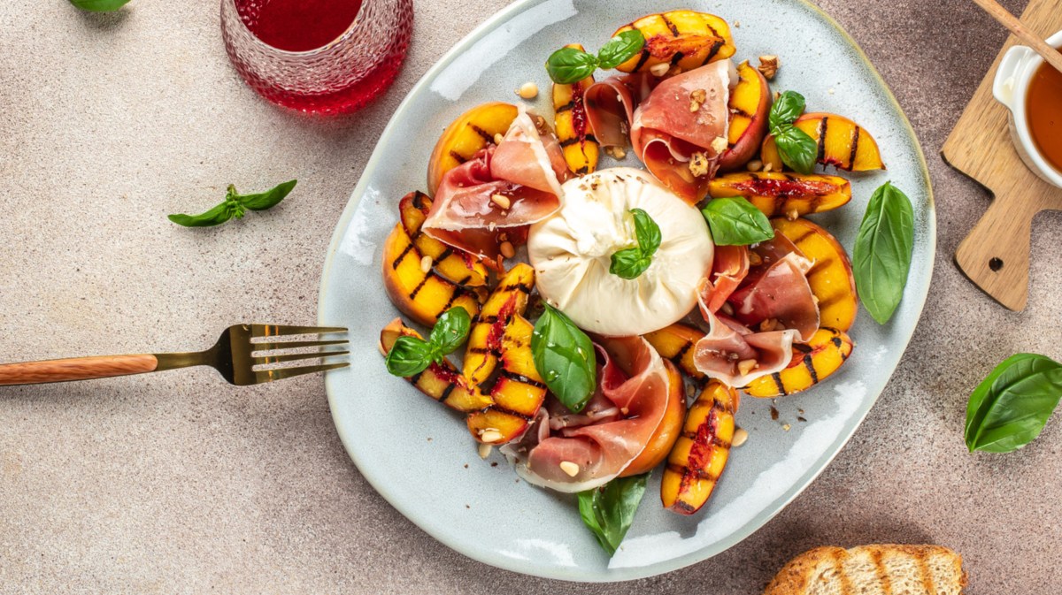 Pfirsich, Burrata, Schinken und Basilikum auf einem Teller. Daneben stehen Getränke und geröstetes Brot.