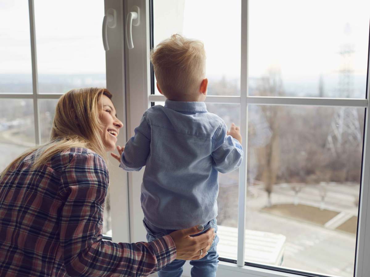 Mutter hält ihren ca. 2-jährigen Sohn fest, während der auf dem Fensterbrett steht und nach draußen schaut.