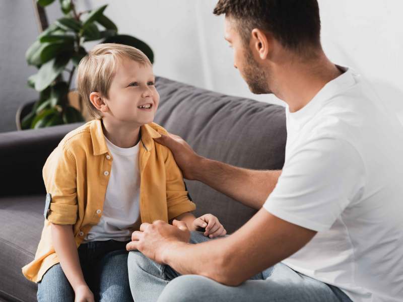 Vater legt seinem ca. 8-jährigen Sohn die Hand auf die Schulter, während beide auf dem Sofa sitzen und quatschen.