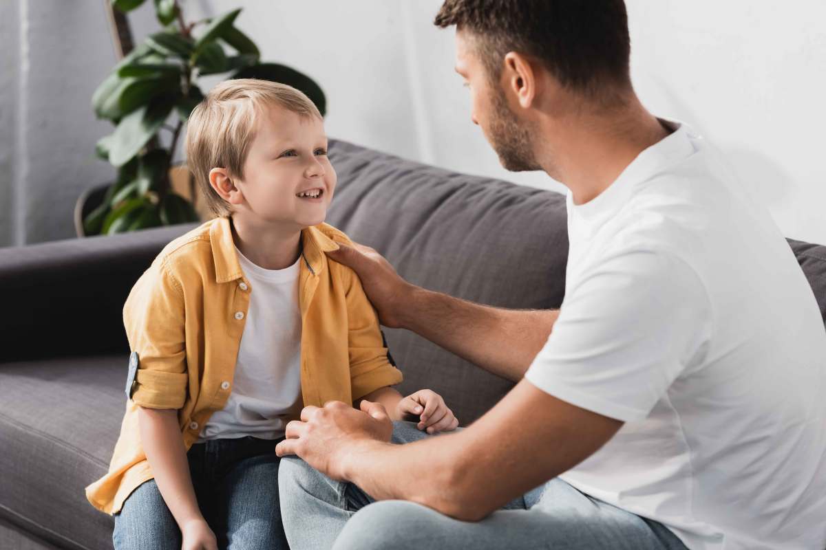 Vater legt seinem ca. 8-jährigen Sohn die Hand auf die Schulter, während beide auf dem Sofa sitzen und quatschen.