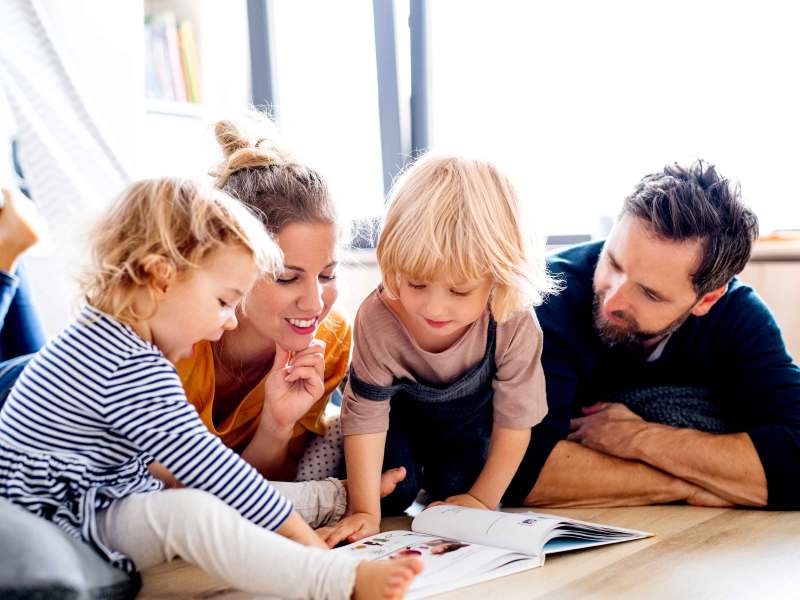 Mutter, Vater und zwei Kinder liegen auf dem Fußboden und blättern gemeinsam in einem Buch.