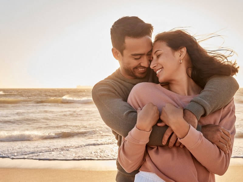 Frau und Mann beim Sonnenuntergang am Strand, die sich kuscheln und verliebt anschauen