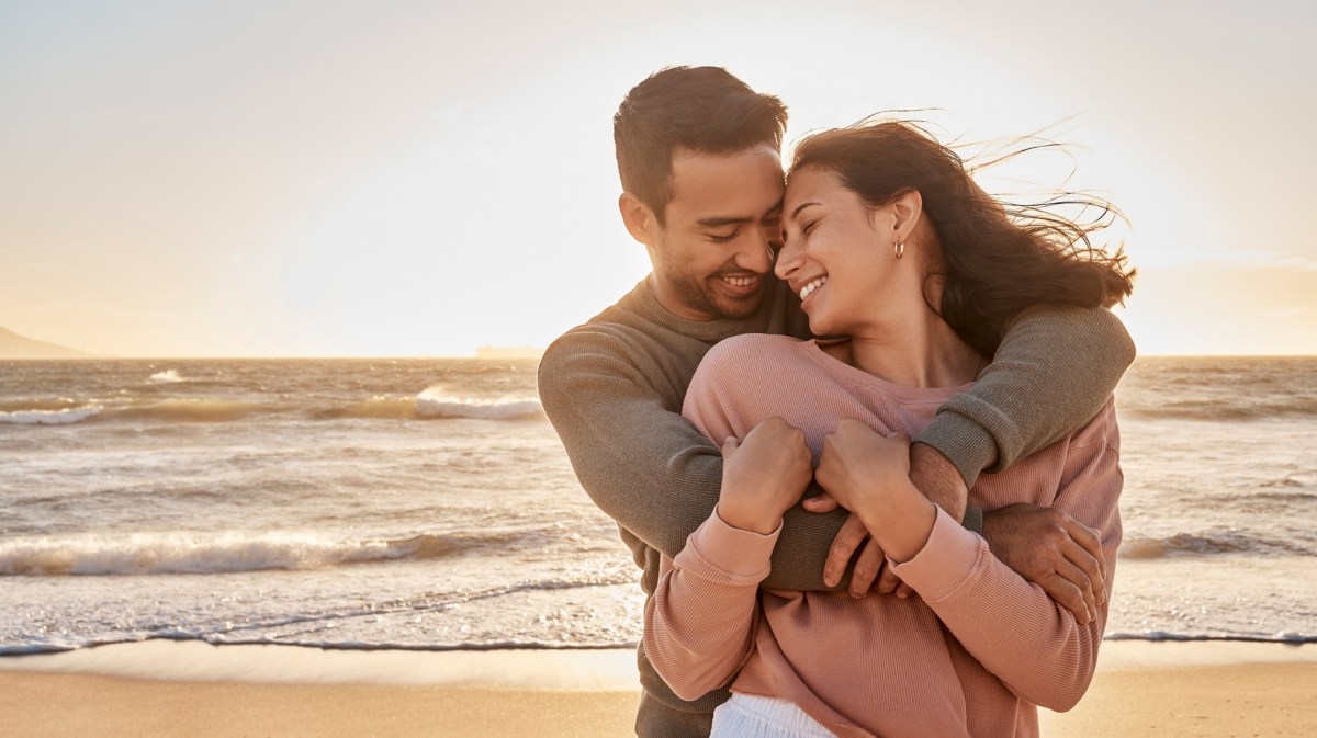 Frau und Mann beim Sonnenuntergang am Strand, die sich kuscheln und verliebt anschauen