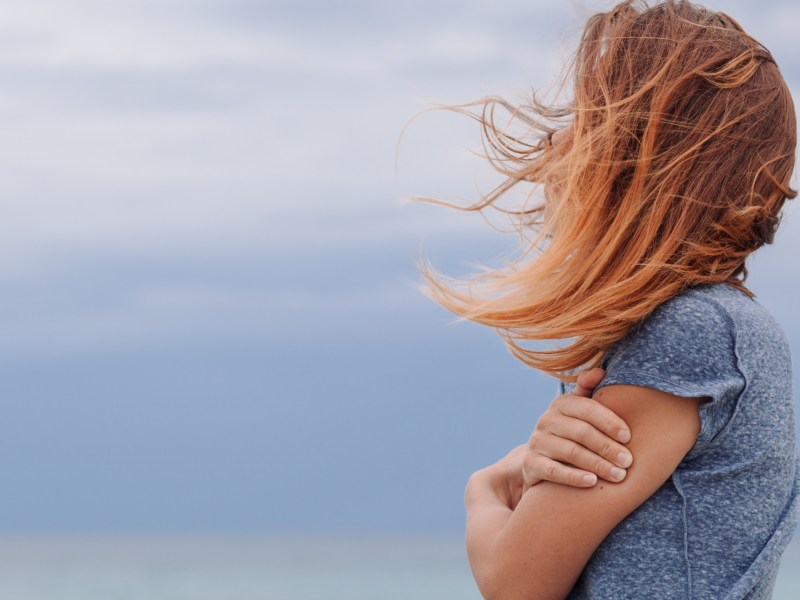 Frau mit blauem Shirt vor blauem Hintergrund umarmt sich selbst.