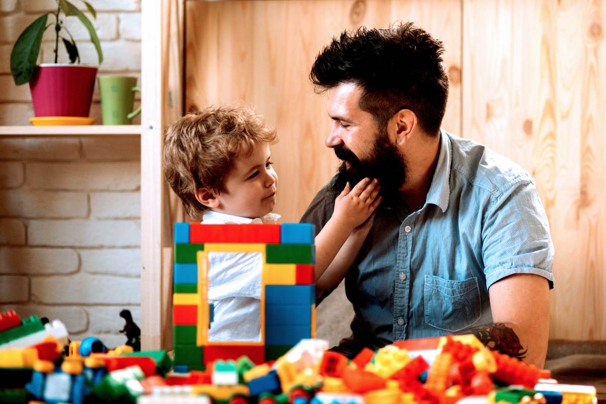 Vater und Sohn sitzen vor einem Stapel Bausteinen. Der Sohn fasst gerade in den Vollbart seines Vaters.