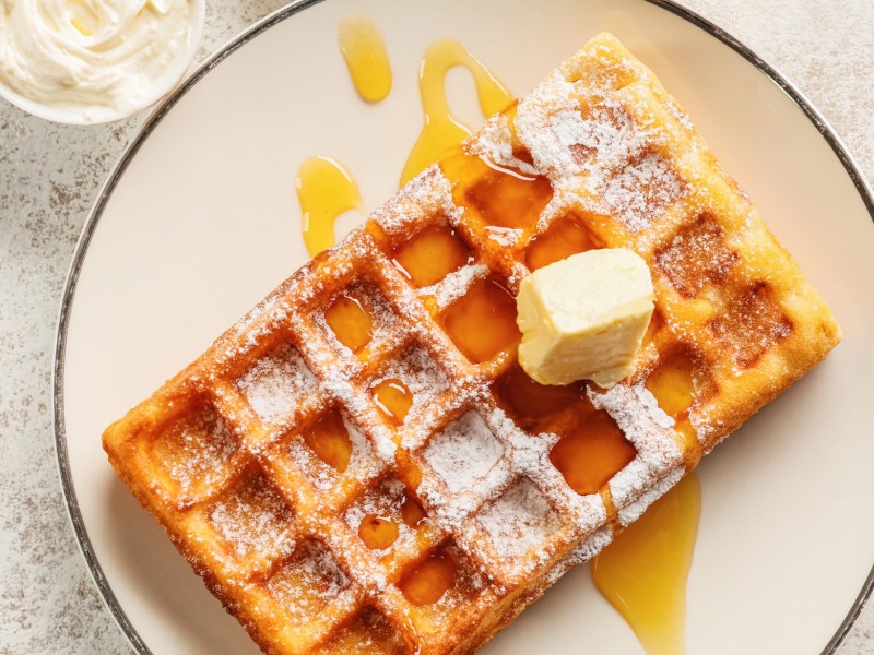 Belgische Waffel auf weißem Teller mit Sirup, Puderzucker und ein Stückchen Butter.