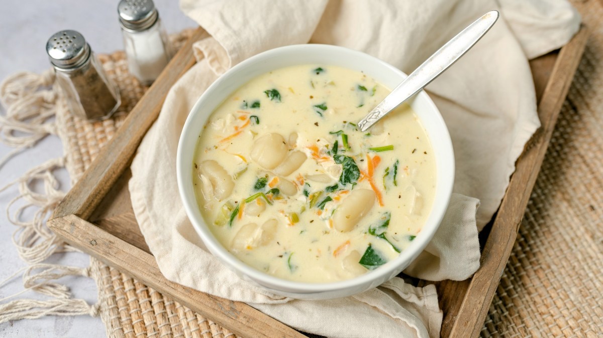 Eine weiße Suppenschale mit der Gnocchi-Suppe auf einem Holzbrett, welches wiederum auf einem weißen Tisch steht. Leicht schräg von oben fotografiert.