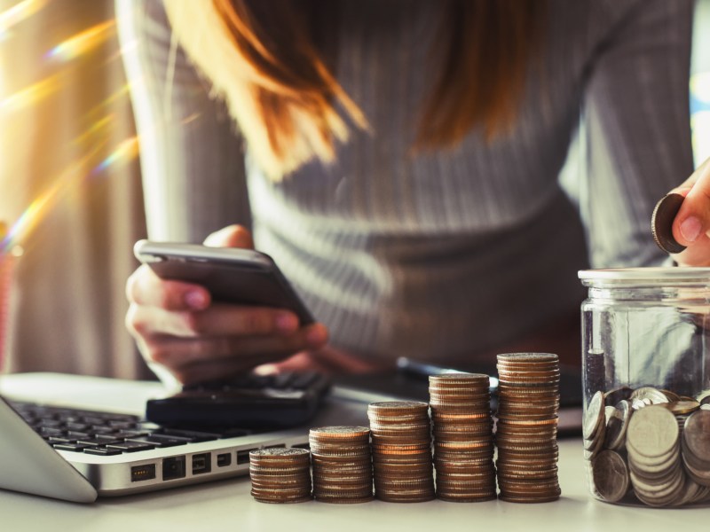 Frau schaut auf Handy und hat mehrere Stapel Münzgeld vor sich auf Tisch liegen.