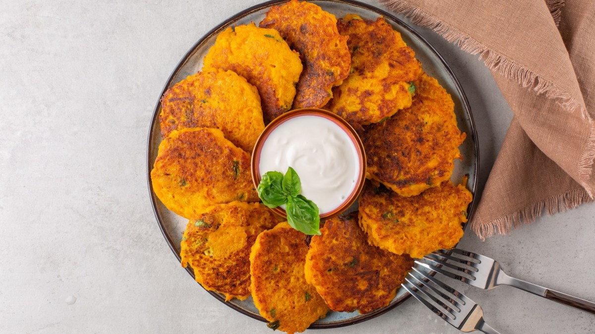 Ein dunkler Teller mit den Kartoffel-Kürbis-Puffern auf hell-grauem Untergrund direkt von oben fotografiert. Rechts unten im Bild ist noch ein braunes Tuch und zwei Gabeln sowie eine kleine Dip-Schale mit weißer Creme in der Mitte.