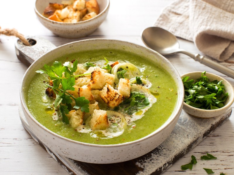 Eine Schüssel Zucchini-Spinat-Suppe mit Croûtons und frischer Petersilie garniert, daneben eine Schüssel mit gehackter Petersilie und eine mit Croûtons, außerdem ein Löffel und ein Geschirrtuch.