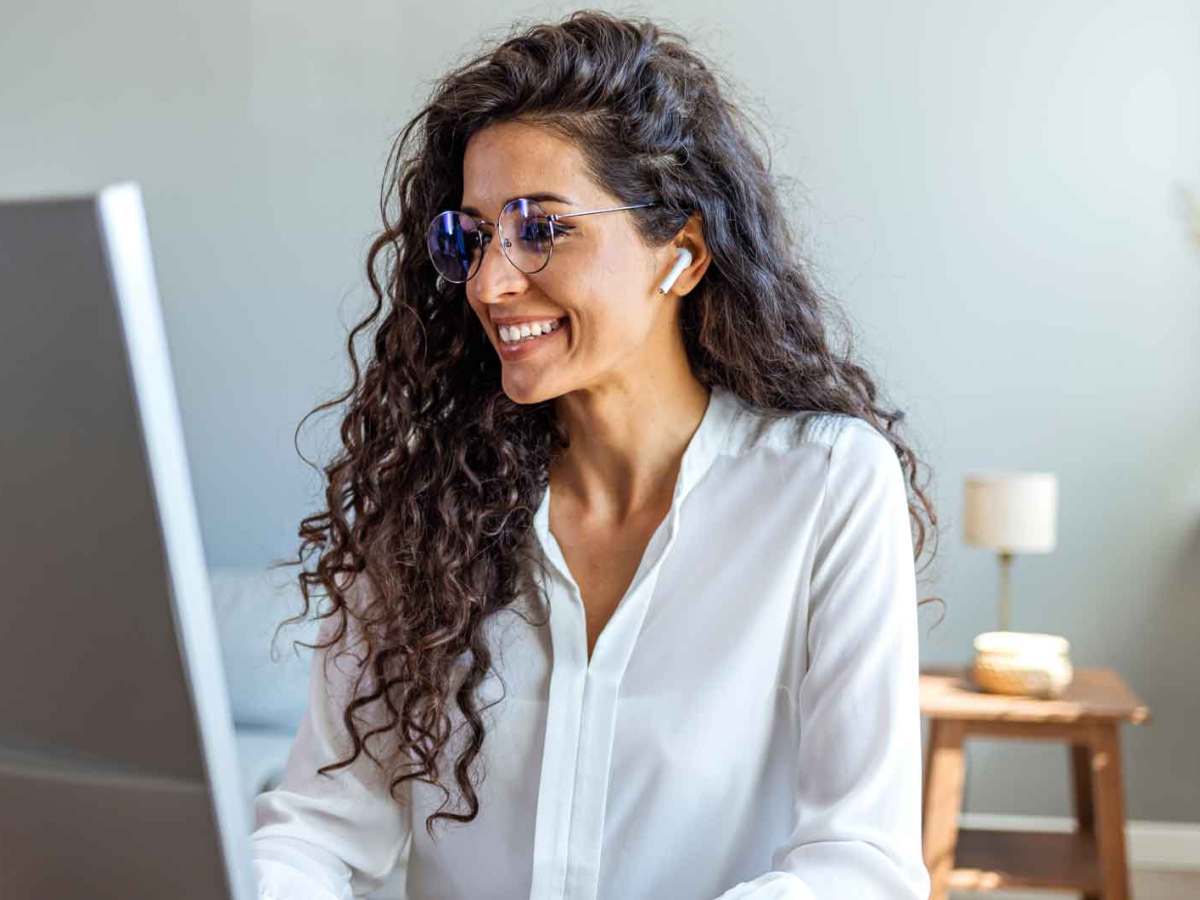 Junge Frau mit dunklen Locken sitzt am Schreibtisch mit Kopfhörern und spricht in einem Online-Meeting mit jemandem.