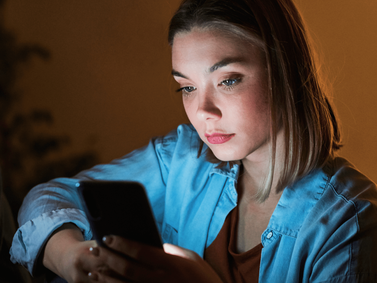 Eine junge Frau mit ernstem Gesichtsausdruck liest auf ihrem Handy, umgeben von ged&auml;mpftem, orangefarbenem Licht.