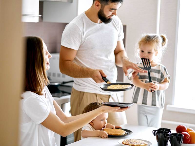 Familie am Küchentisch im alltäglichen Chaos. Der Vater hat Pfannkuchen gemacht. Die Mutter und die beiden Kinder warten ungeduldig aufs Essen.