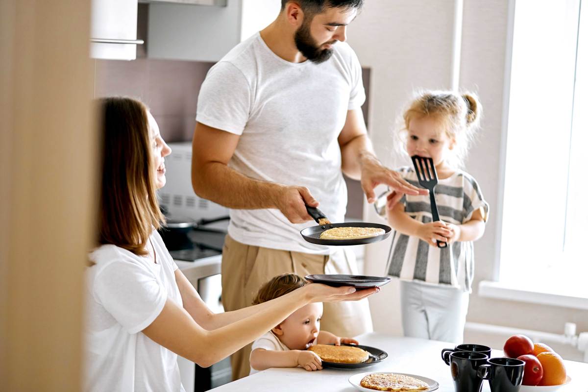 Familie am Küchentisch im alltäglichen Chaos. Der Vater hat Pfannkuchen gemacht. Die Mutter und die beiden Kinder warten ungeduldig aufs Essen.