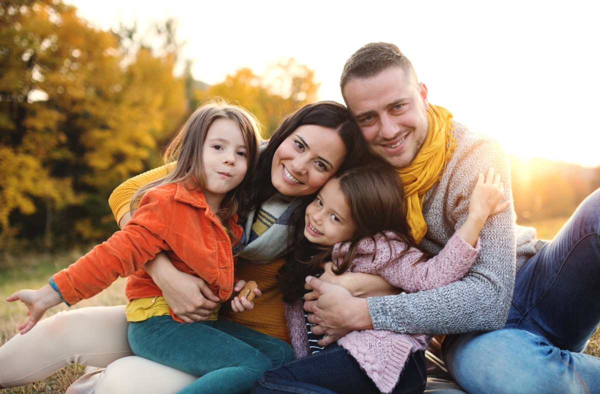 Familie aus Vater, Mutter und zwei Kindern sind in der Natur, sitzen auf einer Decke und lächeln in die Kamera.