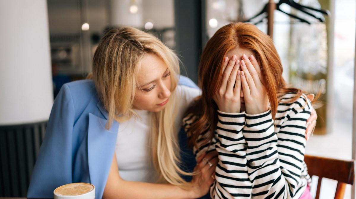 Zwei Frauen in einem Café, die nebeneinander sitzen, während die linke, blonde Frau versucht, die rothaarige, rechte Frau zu trösten