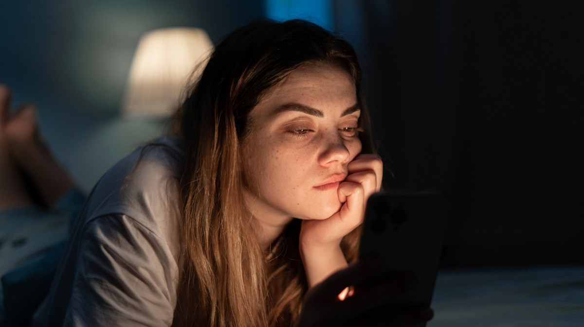 Frau, die gelangweilt in der Dunkelheit auf ihrem Bett liegt und auf ihr Handy Display starrt