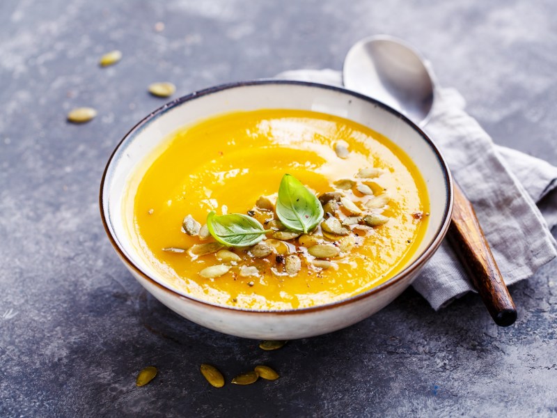 Eine weiße Schüssel Tomaten-Möhren-Suppe garniert mit Kürbiskernen und Basilikum, daneben ein Löffel mit Holzgriff auf einer Serviette.