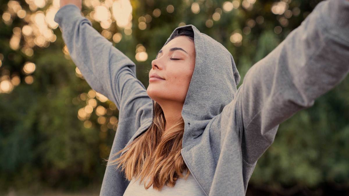 Frau steht morgens draußen in der Natur und atmet tief ein und streckt sich.