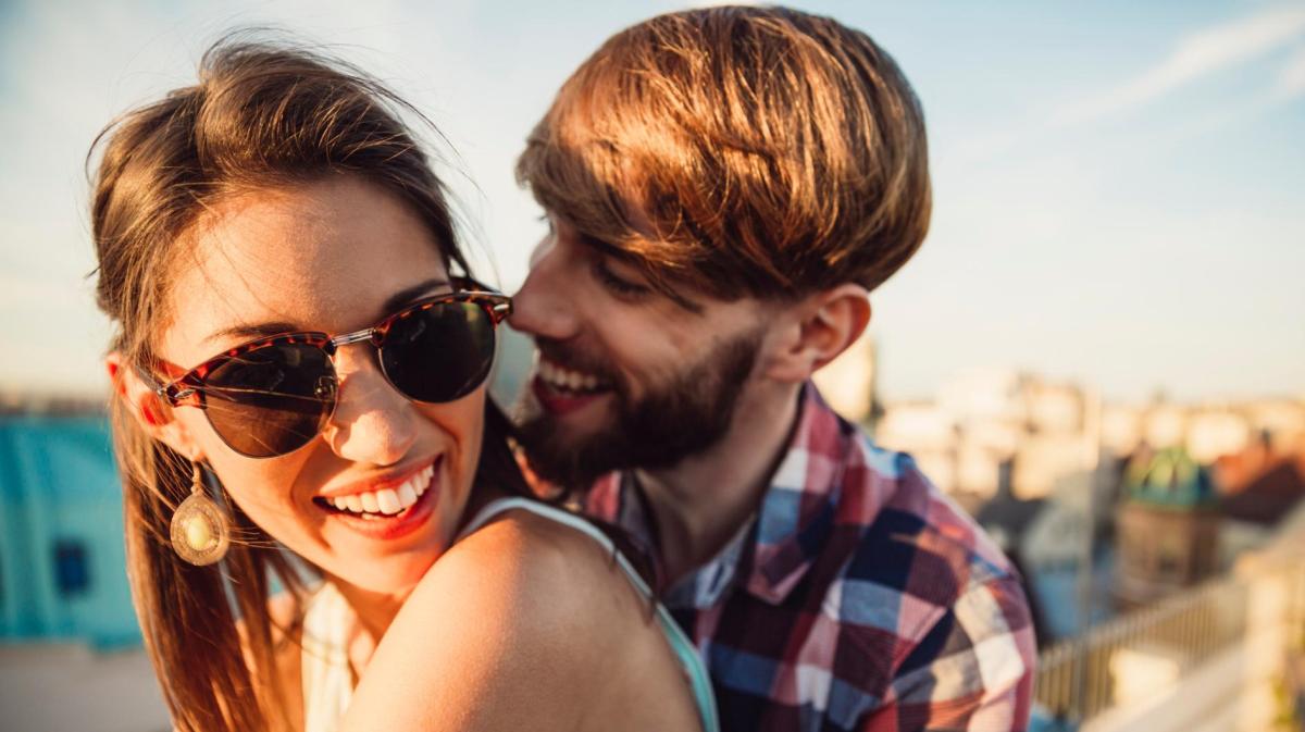 Mann umarmt Frau von hinten. Beide sitzen in der Sonne und sind glücklich.