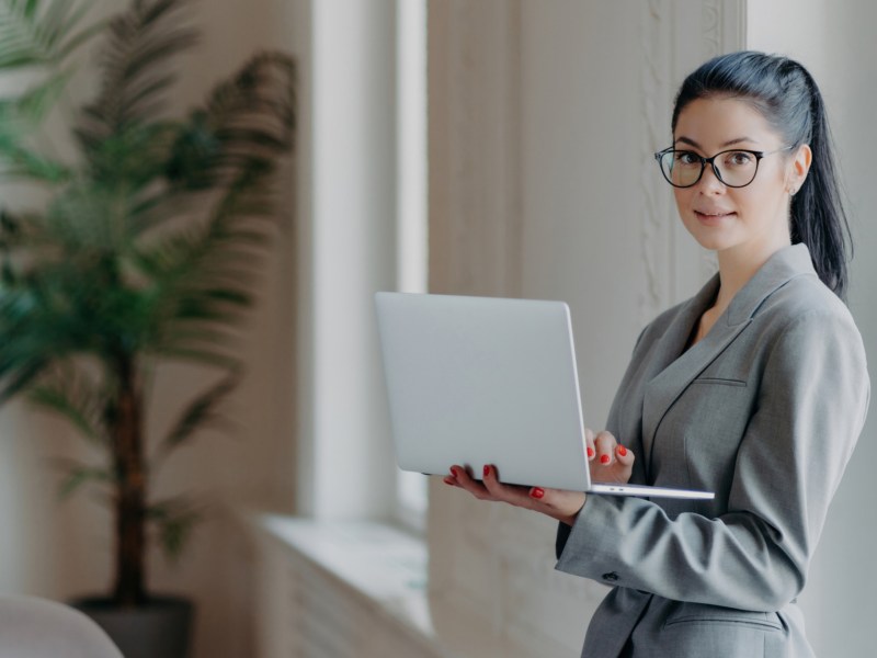 Frau mit Laptop steht auf der Arbeit an einer Wand gelehnt.