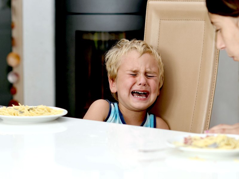 Junge sitzt mit seiner Mutter am Küchentisch und weint fürchterlich. Die Mutter beugt sich leicht zu ihm hinüber.