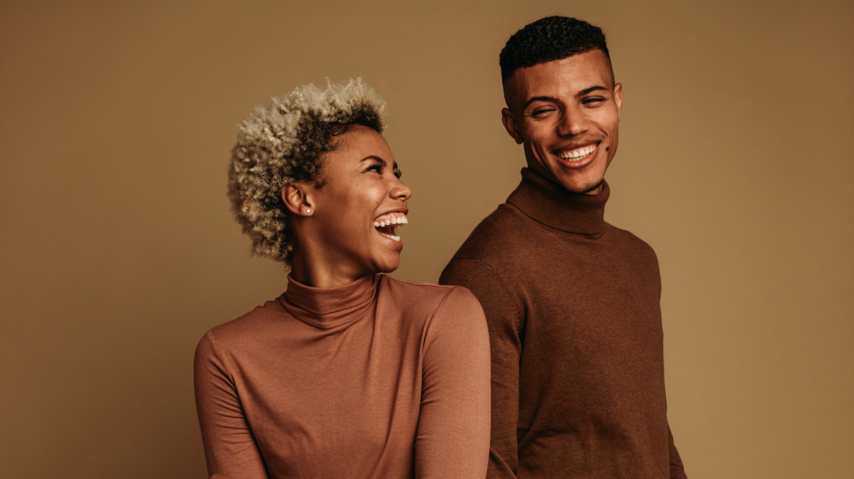 Eine junge Frau mit lockigem blondem Haar lacht herzlich zu einem jungen Mann mit kurzem schwarzen Haar, beide tragen braune Rollkragenpullover.