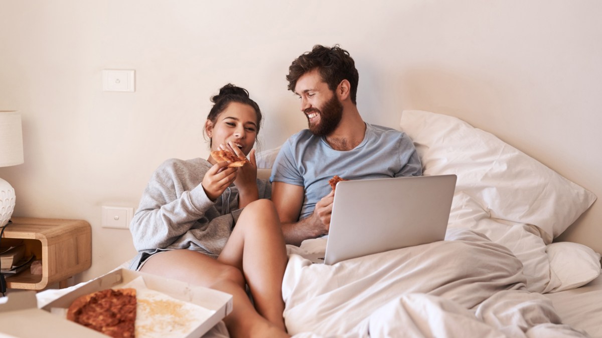 Mann und Frau im Bett, die einen Laptop auf der Bettdecke haben und Pizza essen