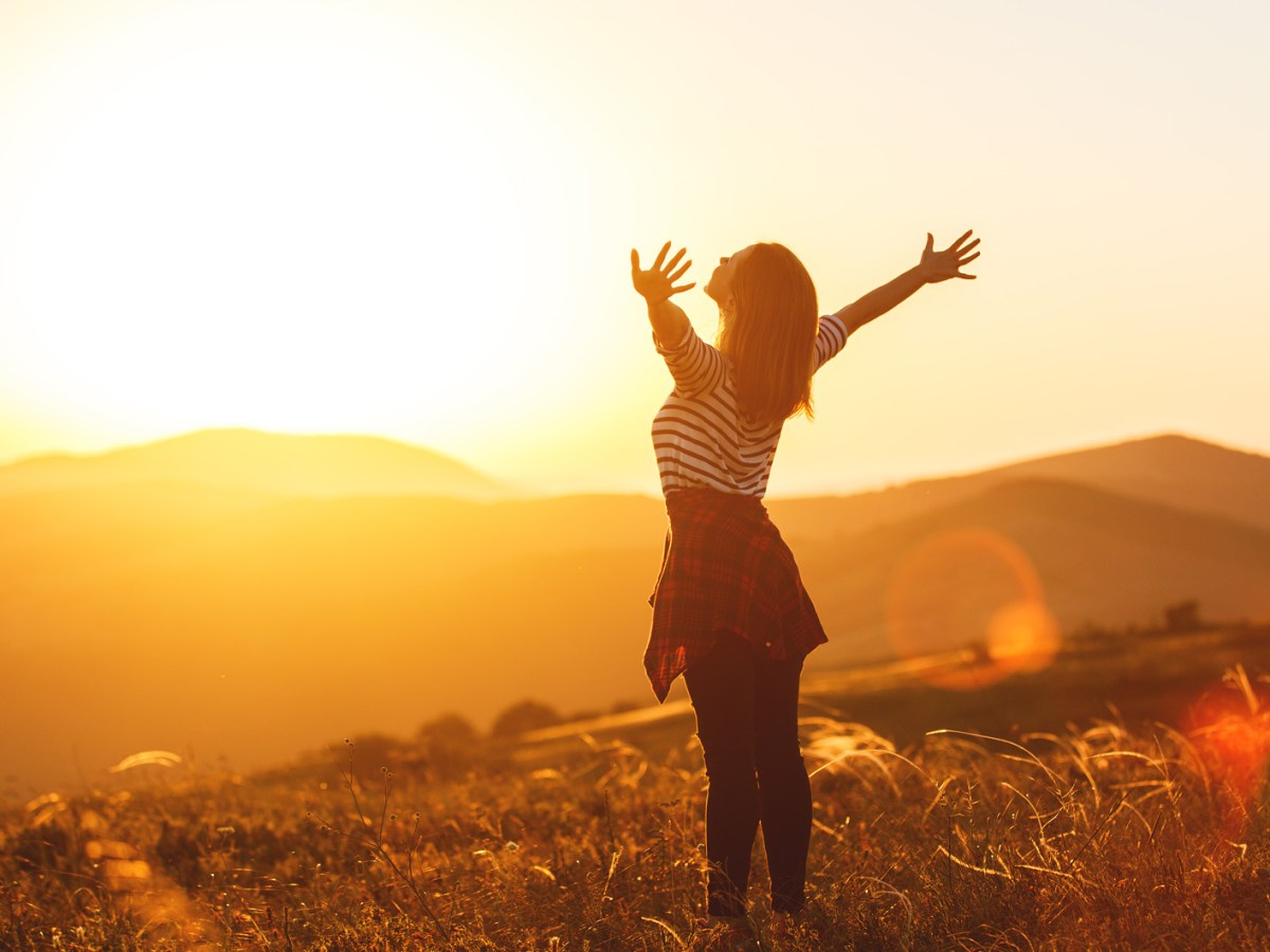 Frau steht vor einer Landschaft bei Sonnenaufgang und streckt die Arme aus.