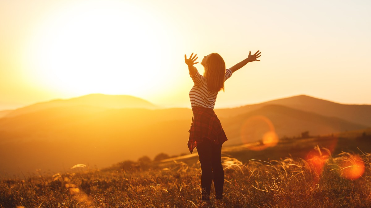 Frau steht vor einer Landschaft bei Sonnenaufgang und streckt die Arme aus.