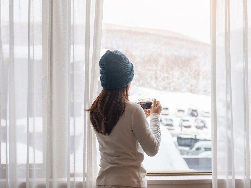 Frau vor dem Fenster, die in eine Winterlandschaft hinausschaut und eine Tasse in der Hand hält