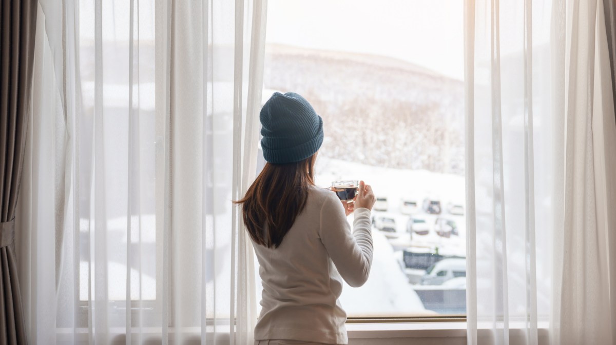 Frau vor dem Fenster, die in eine Winterlandschaft hinausschaut und eine Tasse in der Hand hält