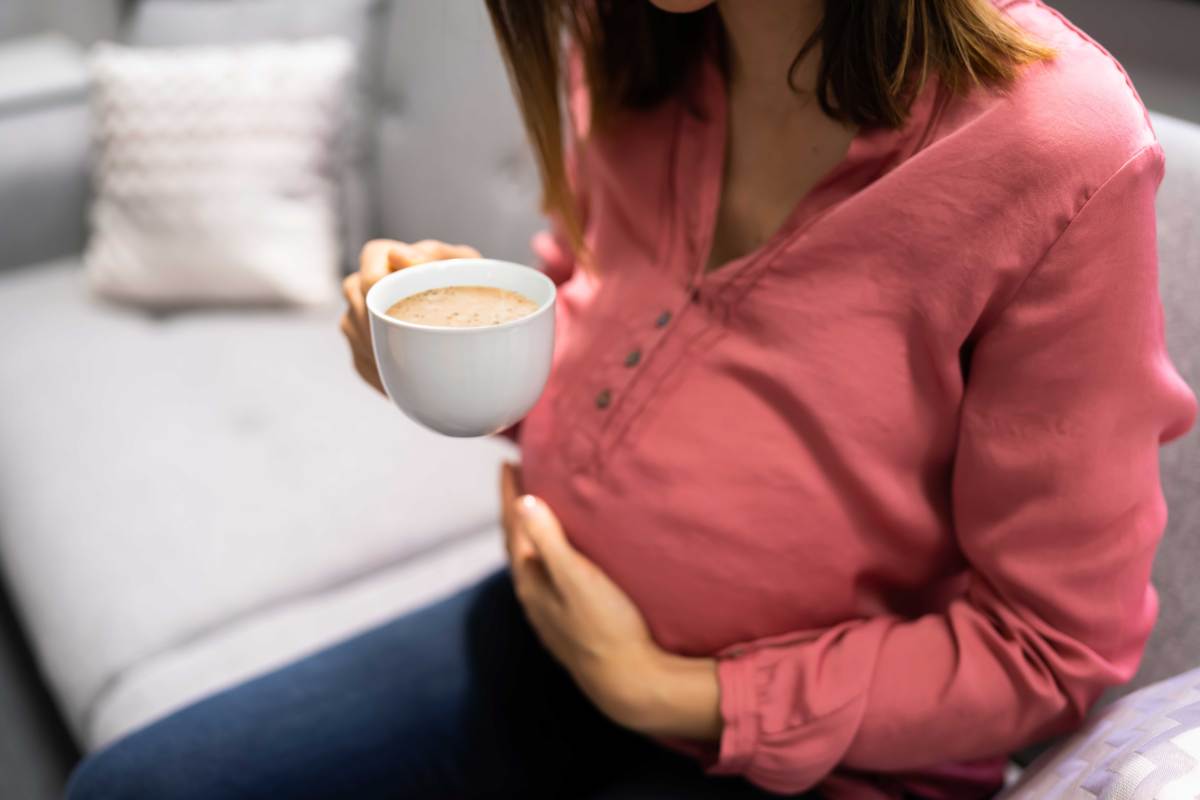 Schwangere Frau sitzt auf dem Sofa, hat eine Hand auf ihren Bauch gelegt und hält in der anderen eine Tasse Kaffee.