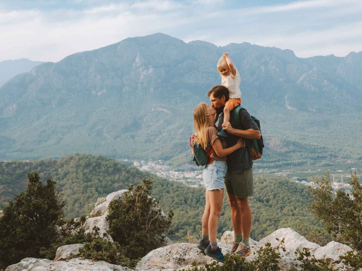 Familie beim Wandern in den Bergen. Sie geben sich einen Kuss, während er das gemeinsame Kind auf den Schultern trägt.