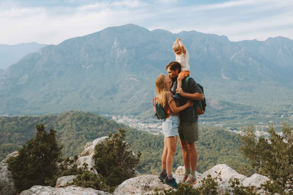 Familie beim Wandern in den Bergen. Sie geben sich einen Kuss, während er das gemeinsame Kind auf den Schultern trägt.