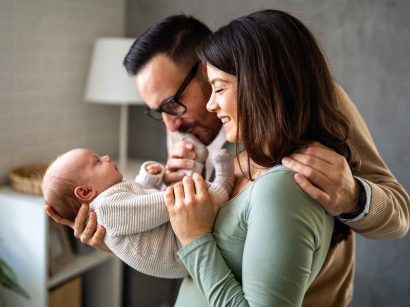 Mutter und Vater halten ihr Neugeborenes in den Armen und schauen es verträumt an.