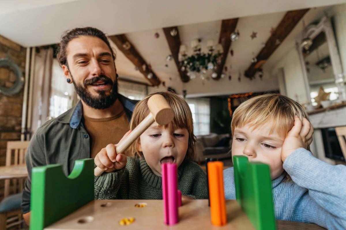 Vater spielt mit seinen beiden Kindern. Eines hat einen Holzhammer in der Hand und versucht, Holznägel in ein Brett zu hämmern.