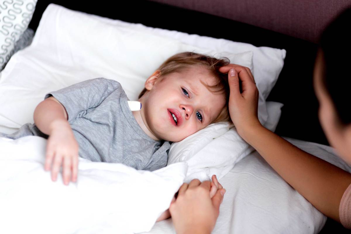 Kleine Junge liegt fiebrig im Bett, das Thermometer unter den Arm geklemmt. Seine Mutter sitzt neben dem Bett und hält seine Hand.