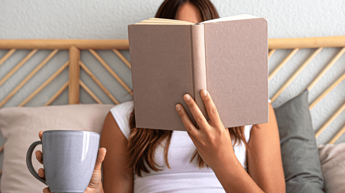 Junge Frau liegt im Bett und liest ein Buch und trinkt einen Tee